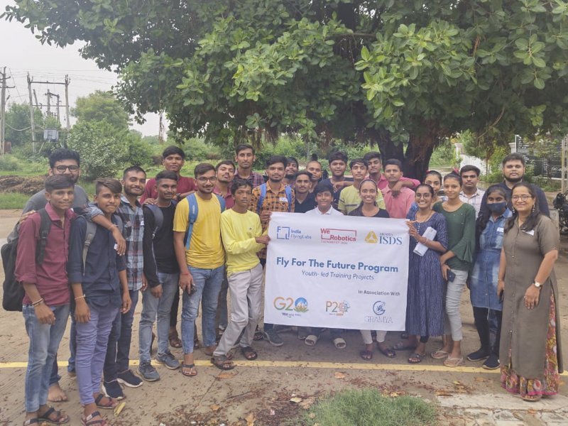 India Flying Labs with participants from the Fly for the Future program. The group stands outside with a tree behind them, and two participants hold up a program banner.