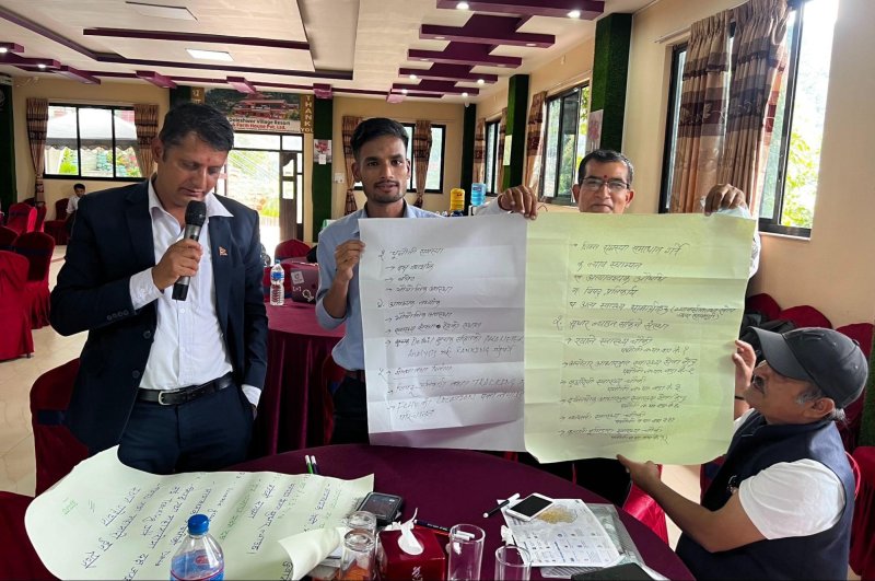 A man speaks into a microphone while reading from a large sheet of paper on a table. Next to him, two men each hold up a sheet of paper with writing on it. A fourth man is seated at the table next to them.