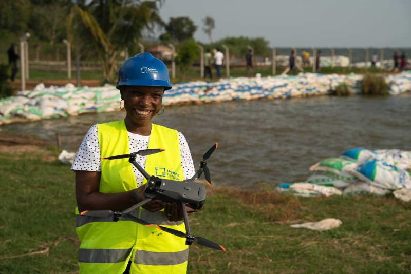 A team member of Uganda Flying Labs smiles as she holds a drone. She is wearing a reflector jacket and Lake Victoria is in the background.