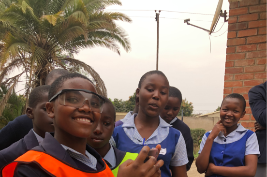 A girl wearing protective eyewear and a reflective jacket over her school uniform smiles at the camera during the youth-led workshop. Her fellow students stand around her.