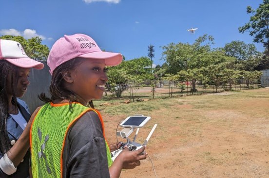 A young woman smiles as she holds a drone controller. Another stands beside her, and ahead of them a drone is in the air.