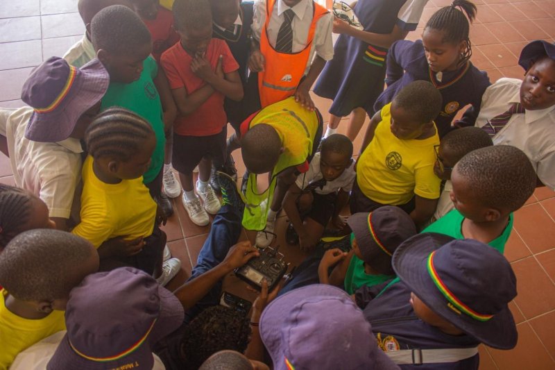 A group of young children huddle around and observe a Zimbabwe Flying Labs team member sitting on the floor holding a controller.