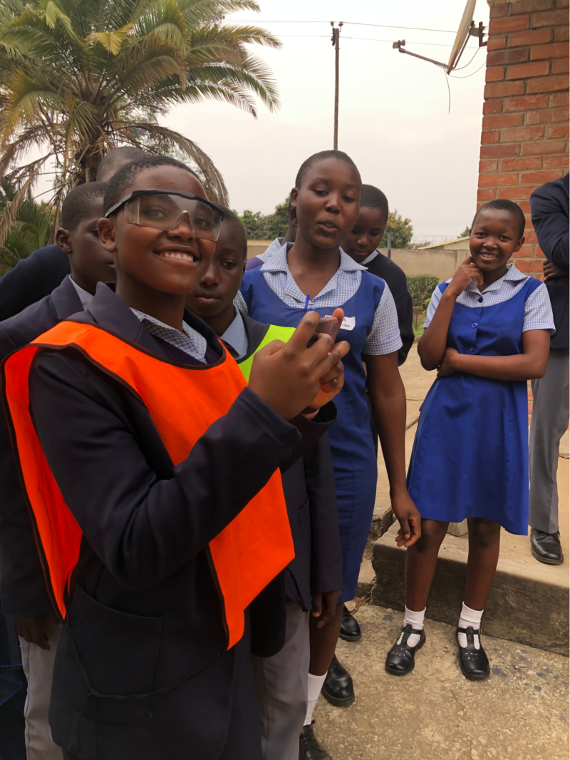 A girl wearing protective eyewear and a reflective jacket over her school uniform smiles at the camera during the youth-led workshop. Her fellow students stand around her.