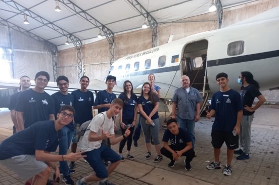 Team members from Brazil Flying Labs take a group photo with students next to a plane in a hangar.