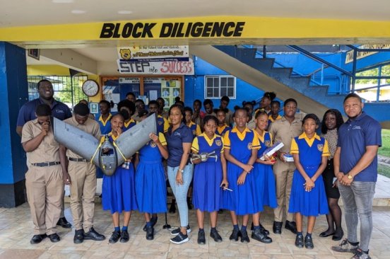 Students in yellow and blue uniform pose for a group photo with the Jamaica Flying Labs team at the National GIS Day celebrations. Some students are holding up a drone.