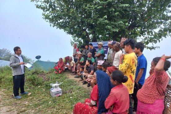 A team member of Nepal Flying Labs stands speaking to a group of community members seated outside in Ramechhap during their TDIA project.