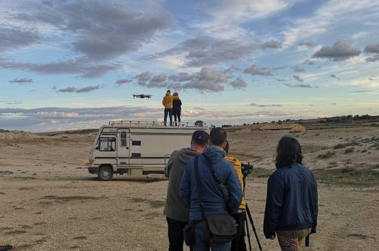 Under a beautiful sky, the Spain Flying Labs team watches a drone in flight in a dry landscape. Their backs are to the camera. Two members stand on top of a van, while four stand on the ground a distance behind the van.