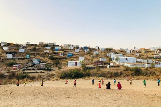 In the foreground children are engaged in a game on a dry field, and in the background is the Brendan Simbwaye A Informal Settlement.