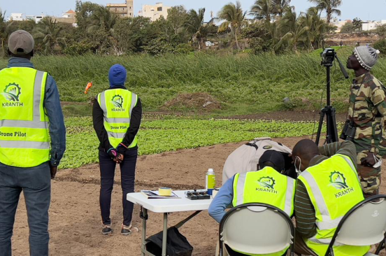 The Senegal Flying Labs team, wearing reflector vests, have their backs to the camera during their mission mapping the Technopole. Three observe the drone in flight while two are seated, hunched over a small table.